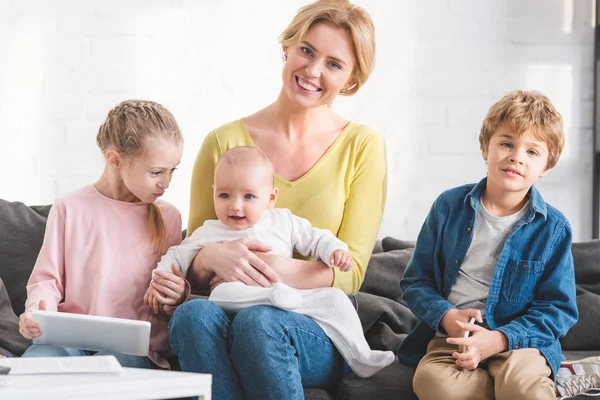Beautiful happy mother smiling at camera while sitting on couch with three adorable kids — Stock Photo