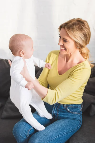 Feliz madre sentada en el sofá y sosteniendo un hermoso bebé en casa - foto de stock