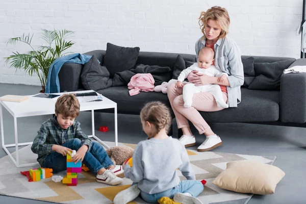 Усталая мать с младенцем сидит на диване и смотрит на братьев и сестер, играющих с игрушками — стоковое фото