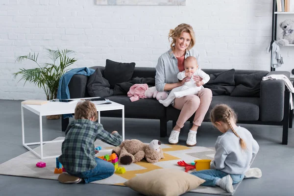 Feliz madre con bebé niño sentado en el sofá y sonriendo a la cámara mientras los hermanos juegan con juguetes - foto de stock