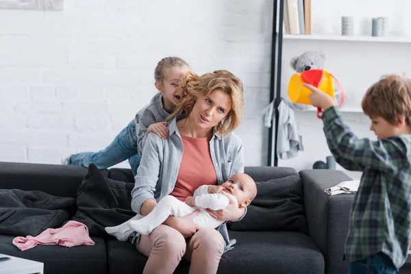 Madre stanca con bambino seduto sul divano mentre i bambini cattivi giocano a casa — Foto stock