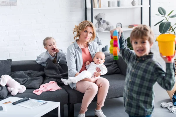 Tired mother with infant child sitting on couch while naughty siblings playing at home — Stock Photo