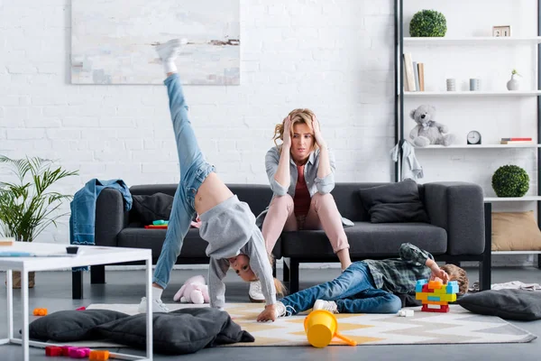 Müde Mutter sitzt auf der Couch und schaut freche Kinder an, die zu Hause spielen — Stockfoto