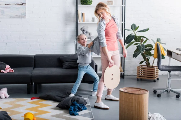 Traviesa hija sosteniendo cárdigan de madre cansada limpieza en casa - foto de stock