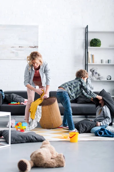 Madre cansada poniendo juguetes en la cesta mientras los niños traviesos pelean con almohadas - foto de stock