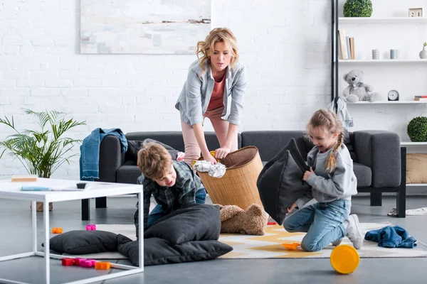 Emocional cansado mãe colocando brinquedos na cesta enquanto crianças safadas lutando com travesseiros — Fotografia de Stock