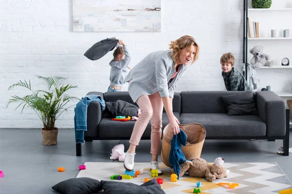 Mère fatiguée émotionnelle mettre des jouets dans le panier tandis que les enfants coquins jouer derrière — Photo de stock