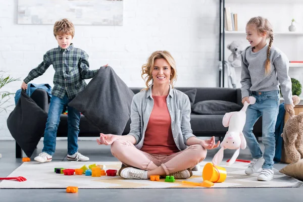 Madre meditando in posizione di loto e sorridendo alla fotocamera mentre i bambini giocano a casa — Foto stock