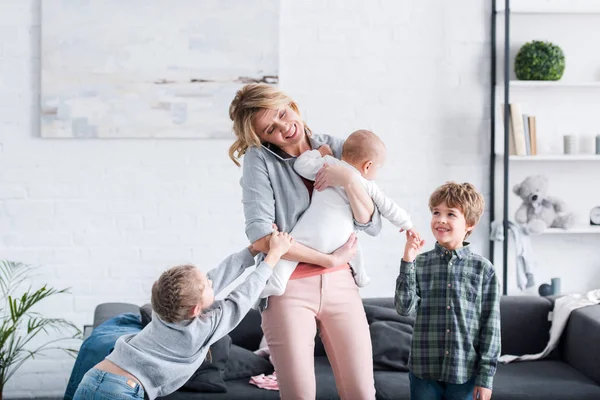 Mère épuisée parlant par smartphone et tenant l'enfant en bas âge tandis que les frères et sœurs coquins jouent dans la chambre — Photo de stock