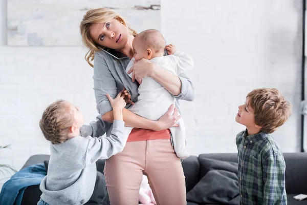 Madre cansada hablando por teléfono inteligente y sosteniendo al bebé mientras los hermanos están de pie en la habitación - foto de stock