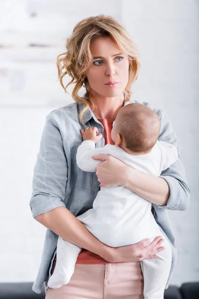 Задумчивая мать держит младенца ребенка и смотрит в сторону дома — стоковое фото