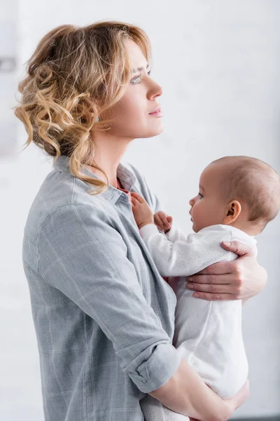 Seitenansicht einer nachdenklichen Mutter, die ihr entzückendes Kleinkind hält und zu Hause wegsieht — Stockfoto