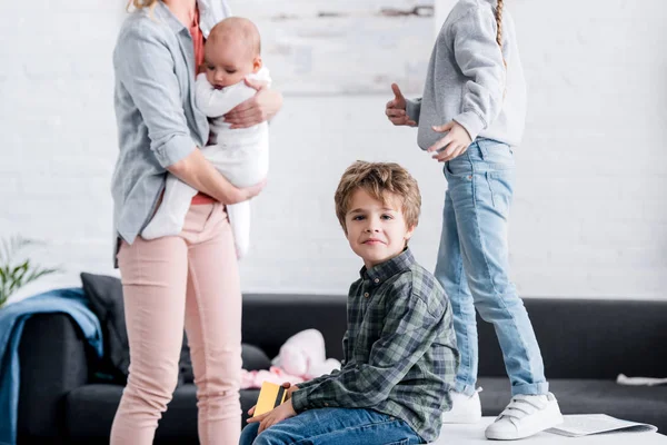 Niño pequeño sosteniendo la tarjeta de crédito y mirando a la cámara mientras la hermana y la madre con el niño de pie detrás - foto de stock