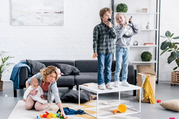 Erschöpfte Mutter mit Säugling auf dem Boden sitzend, während freche Kinder zu Hause spielen — Stockfoto