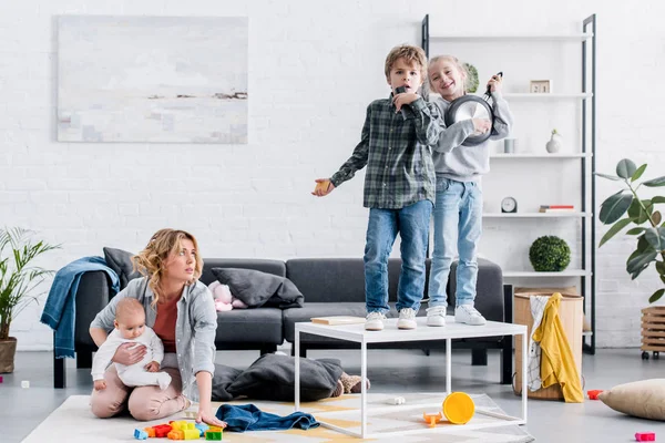 Madre cansada sosteniendo niño bebé mientras niños traviesos jugando en casa - foto de stock