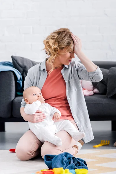 Exhausted mother holding infant child and sitting with hand on forehead on floor — Stock Photo