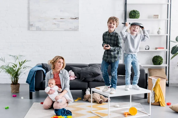 Exhausted mother holding infant child while naughty kids playing at home — Stock Photo