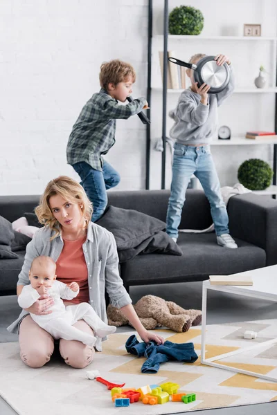 Esausto madre tenendo bambino neonato e guardando la fotocamera mentre i bambini cattivi giocare a casa — Foto stock
