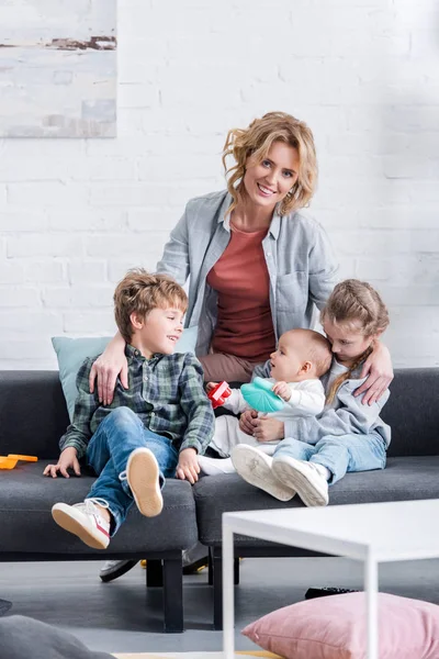 Felice madre sorridente alla macchina fotografica mentre trascorre del tempo con tre adorabili bambini a casa — Foto stock