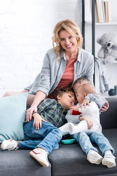 Hermosa madre feliz sonriendo a la cámara mientras tres niños adorables abrazándose en el sofá - foto de stock