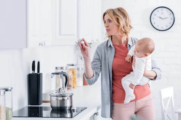 Madre sosteniendo niño adorable bebé y cocinar en la cocina - foto de stock