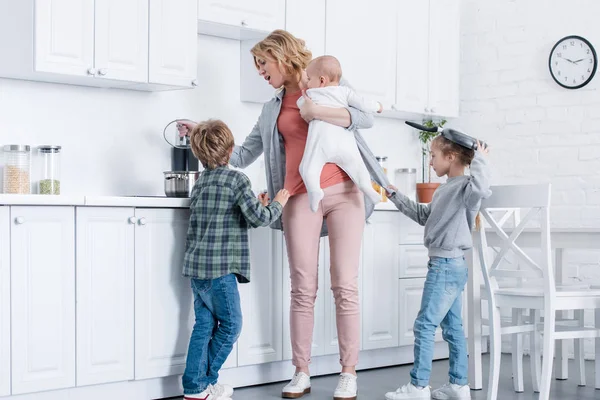 Tired mother with infant kid cooking while naughty children playing in kitchen — Stock Photo