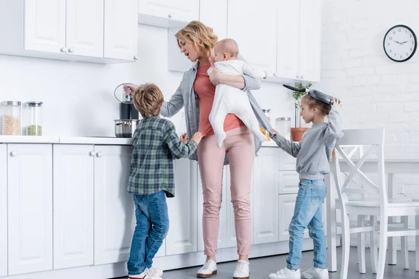 Madre cansada con niño bebé cocinando mientras niños traviesos jugando en la cocina - foto de stock