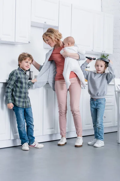 Mère épuisée avec enfant en bas âge cuisine tandis que les enfants coquins jouent dans la cuisine — Photo de stock