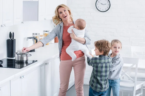 Madre agotada con niño pequeño cocinando y mirando hacia arriba mientras niños traviesos jugando en la cocina - foto de stock