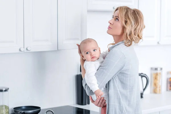 Уставшая мать держит младенца ребенка и глядя вверх на кухне — стоковое фото