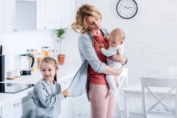 Madre cansada sosteniendo niño bebé mientras hija traviesa sosteniendo su chaqueta en la cocina - foto de stock