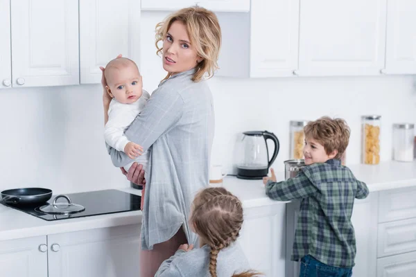 Madre cansada sosteniendo niño bebé y mirando a la cámara mientras niños traviesos jugando en la cocina — Stock Photo