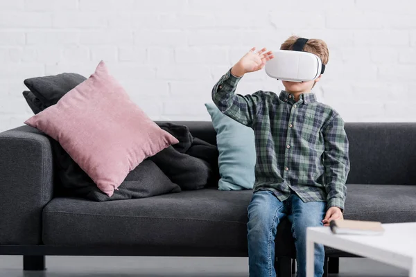 Очаровательный мальчик сидит на диване и использует гарнитуру виртуальной реальности дома — стоковое фото
