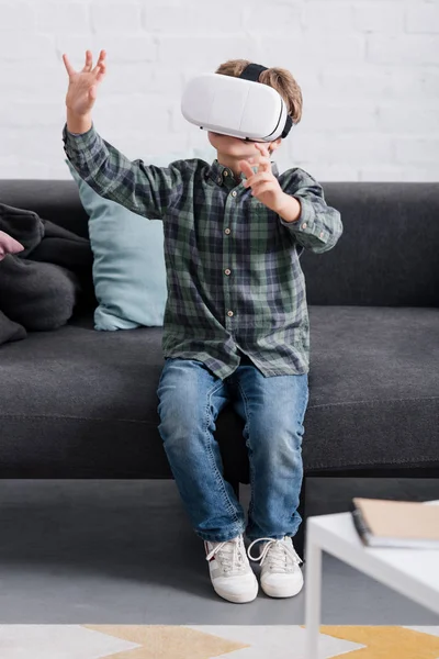 Ребенок сидит на диване и с помощью гарнитуры виртуальной реальности дома — стоковое фото