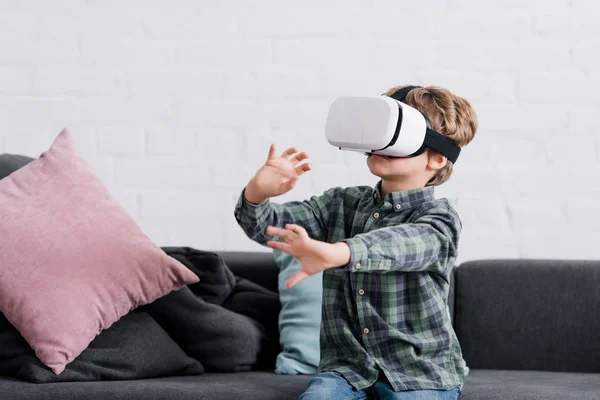 Menino bonito sentado no sofá e usando fone de ouvido realidade virtual — Fotografia de Stock