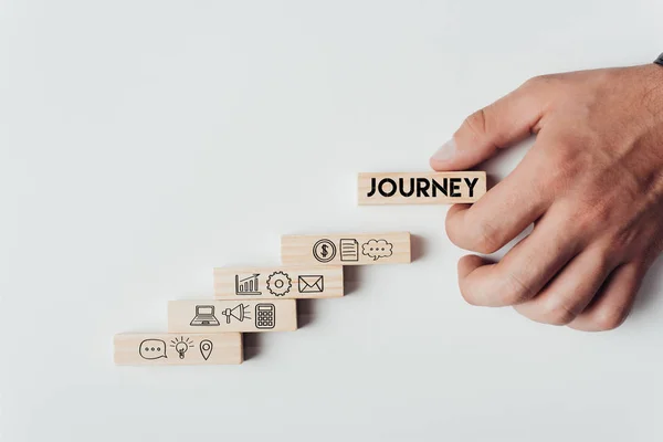 Cropped view of man holding wooden block with word 'journey' on top of wooden bricks with icons isolated on white — Stock Photo