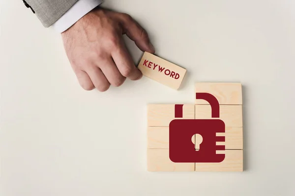 Vista parcial del hombre sosteniendo ladrillo con letras de 'palabra clave' sobre bloques de madera con icono de bloqueo aislado en blanco - foto de stock