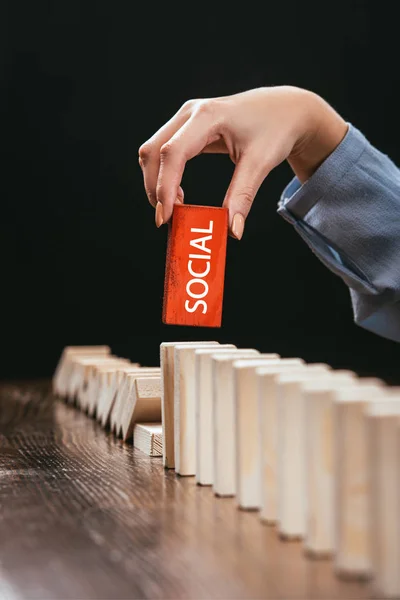Partial view of woman picking red wooden brick with word 'social' from row of blocks on desk isolated on black — Stock Photo