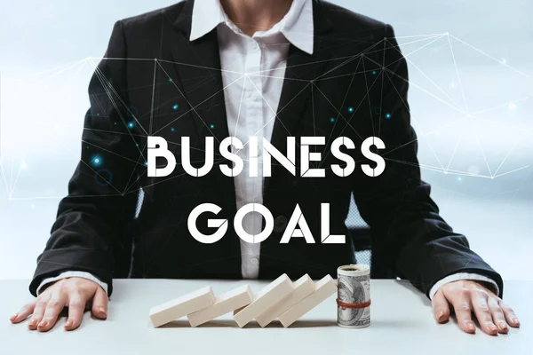 Cropped view of businesswoman with wooden blocks and money roll on table and 'business goal' lettering on foreground — Stock Photo