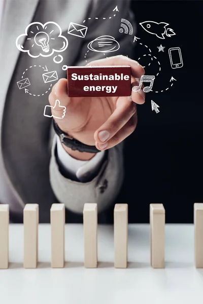 Partial view of man holding red brick with words 'sustainable energy' isolated on black, icons on foreground — Stock Photo