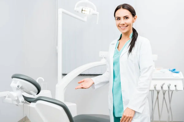 Sorridente dentista feminino mostrando cadeira na clínica odontológica — Fotografia de Stock