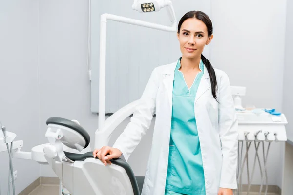 Attraente dentista in piedi vicino sedia in clinica dentale — Foto stock