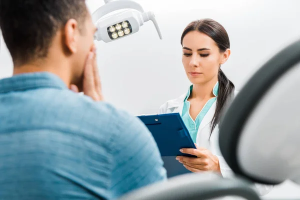 Enfoque selectivo del dentista femenino mirando el portapapeles y de pie cerca de un paciente afroamericano con dolor de muelas - foto de stock