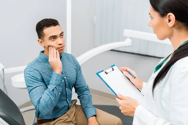 Female dentist standing with clipboard and looking at handsome african american patient having toothache — Stock Photo