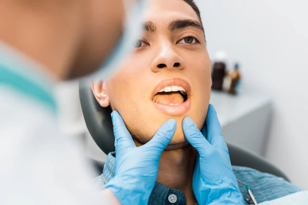 Messa a fuoco selettiva di mani dentiste femminili in guanti di lattice toccare il viso di bello uomo africano americano con bocca aperta — Foto stock