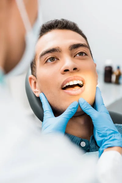 Foco seletivo das mãos do dentista em luvas de látex tocando o rosto do homem afro-americano com boca aberta — Fotografia de Stock