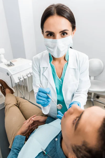 Estomatóloga en guantes de látex y mascarilla examinando paciente afroamericano en silla - foto de stock