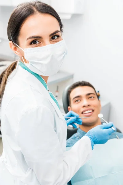 Dentista feminina segurando instrumentos médicos nas mãos e de pé em máscara perto de paciente afro-americano — Fotografia de Stock