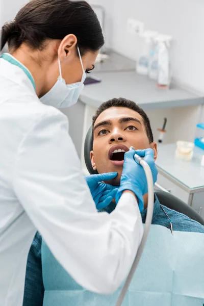 Dentista hembra perforando dientes de paciente afroamericano en silla - foto de stock