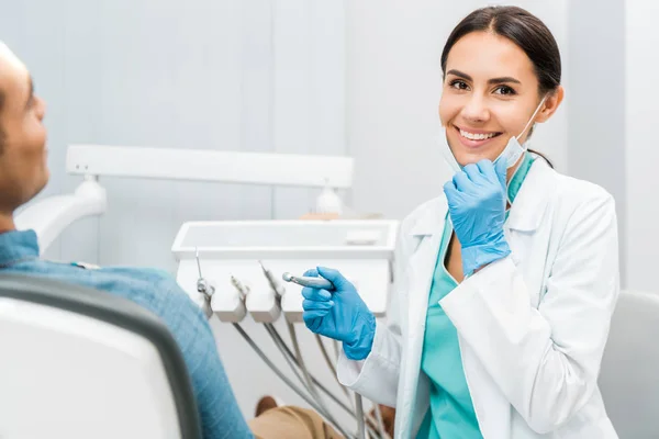 Веселая женщина-дантист держит дрель и улыбается рядом с пациентом — стоковое фото